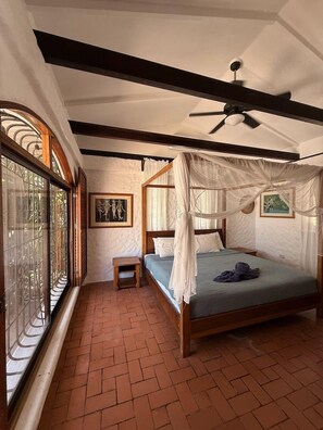 A bedroom with a four-poster bed, a ceiling fan, a small bedside table, and a window with a decorative grille.