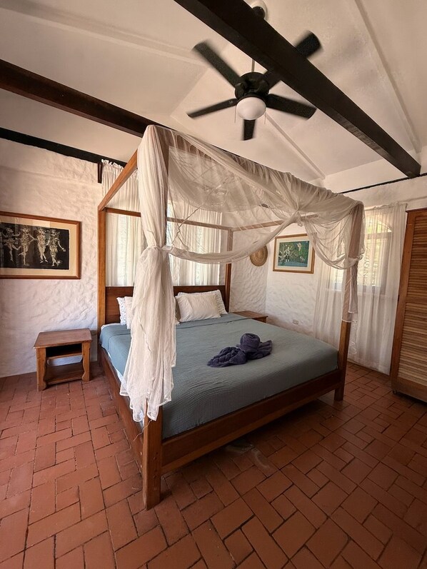 A bedroom with a four-poster bed, a ceiling fan, a small wooden bench, and a brick floor.