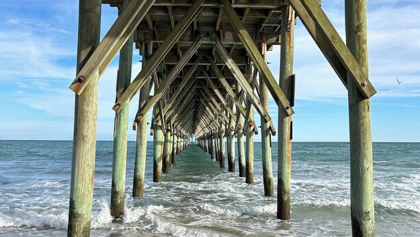 A wooden pier extending into the ocean with waves lapping at its base. - FALL SPECIAL PRICING Beautifully Remodeled Surf City Oceanfront on the beach (Surf City)