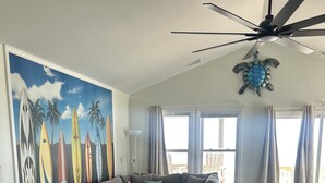 A living room with a sectional sofa, a ceiling fan, a mural of surfboards, and a wooden floor. - FALL SPECIAL PRICING Beautifully Remodeled Surf City Oceanfront on the beach (Surf City)