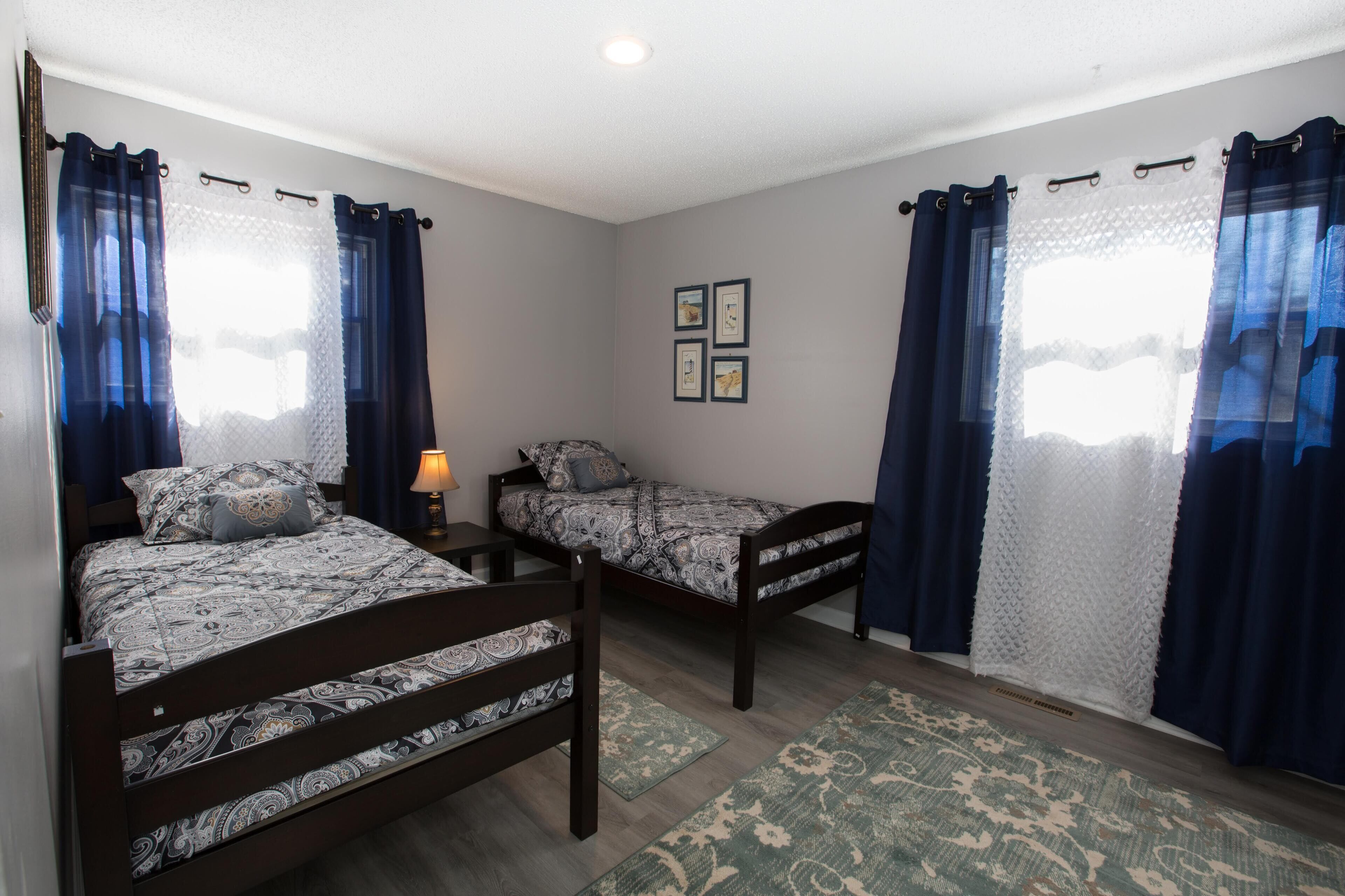 Una habitación con dos camas, cada una con ropa de cama estampada, una mesita de noche pequeña con lámpara, y un cuadro colgado en la pared.