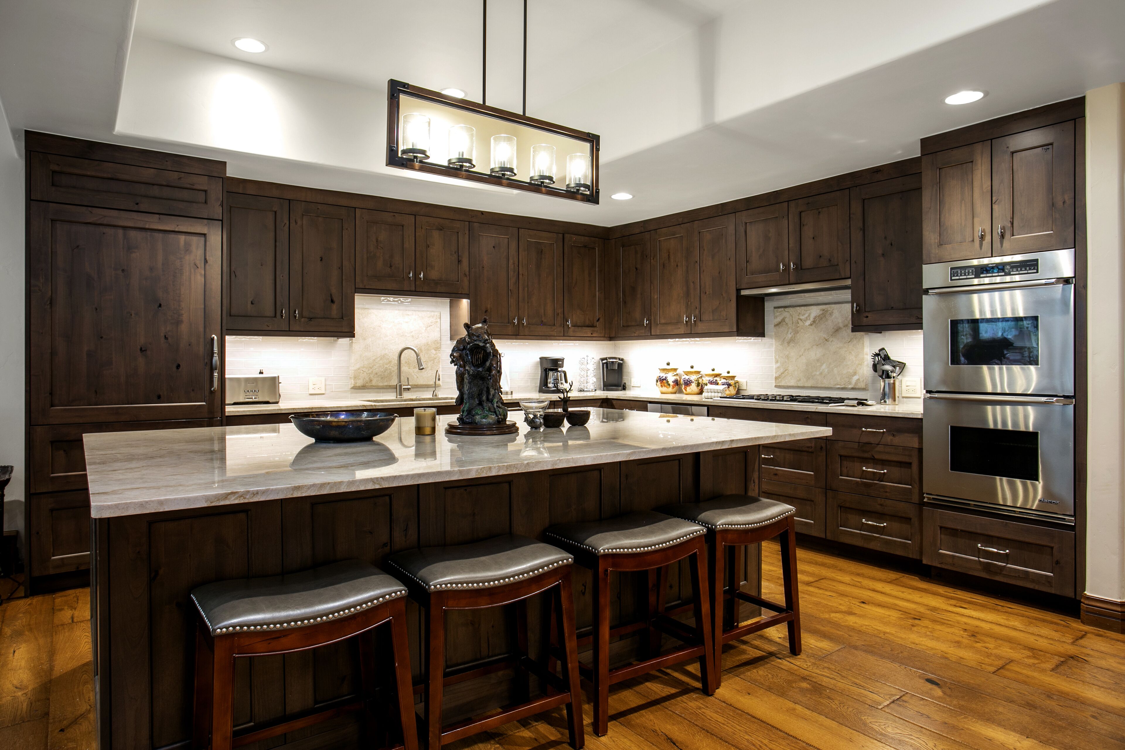 A modern kitchen with dark wooden cabinets, a large island with bar stools, stainless steel appliances, and a pendant light fixture.