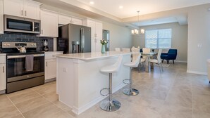Uma cozinha moderna com eletrodomésticos de aço inoxidável, uma ilha central com banquetas e uma área de jantar com uma cadeira azul.