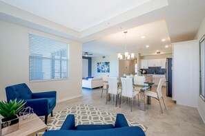 Una sala moderna con un sofá azul, un comedor con mesa de vidrio y una cocina al fondo.