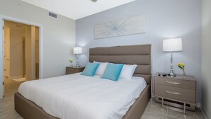 Quarto com uma cama grande, duas mesas de cabeceira, um ventilador de teto, uma obra de arte na parede e uma porta que dá para outro cômodo.