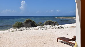 Une vue sur la plage, avec un rivage sablonneux, une chaise longue en bois et un promontoire rocheux couvert de végétation.