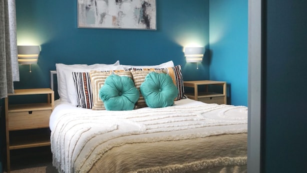 A bedroom with a bed, two bedside tables, a wall-mounted abstract painting, and two wall-mounted lamps. - The Village House (Belfast)