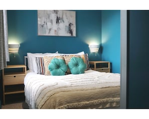 A bedroom with a bed, two bedside tables, a wall-mounted abstract painting, and two wall-mounted lamps. - The Village House (Belfast)
