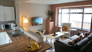 A modern kitchen with a wooden countertop, a built-in oven, and a sink. There is a dining area with a table and chairs, and a sofa with patterned cushions. A TV is mounted on the wall, and a plant is on a shelf. - The Village House (Belfast)