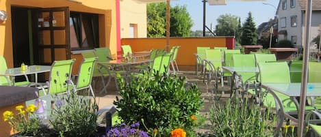 An outdoor seating area with tables and chairs, a large umbrella, and potted plants with flowers.