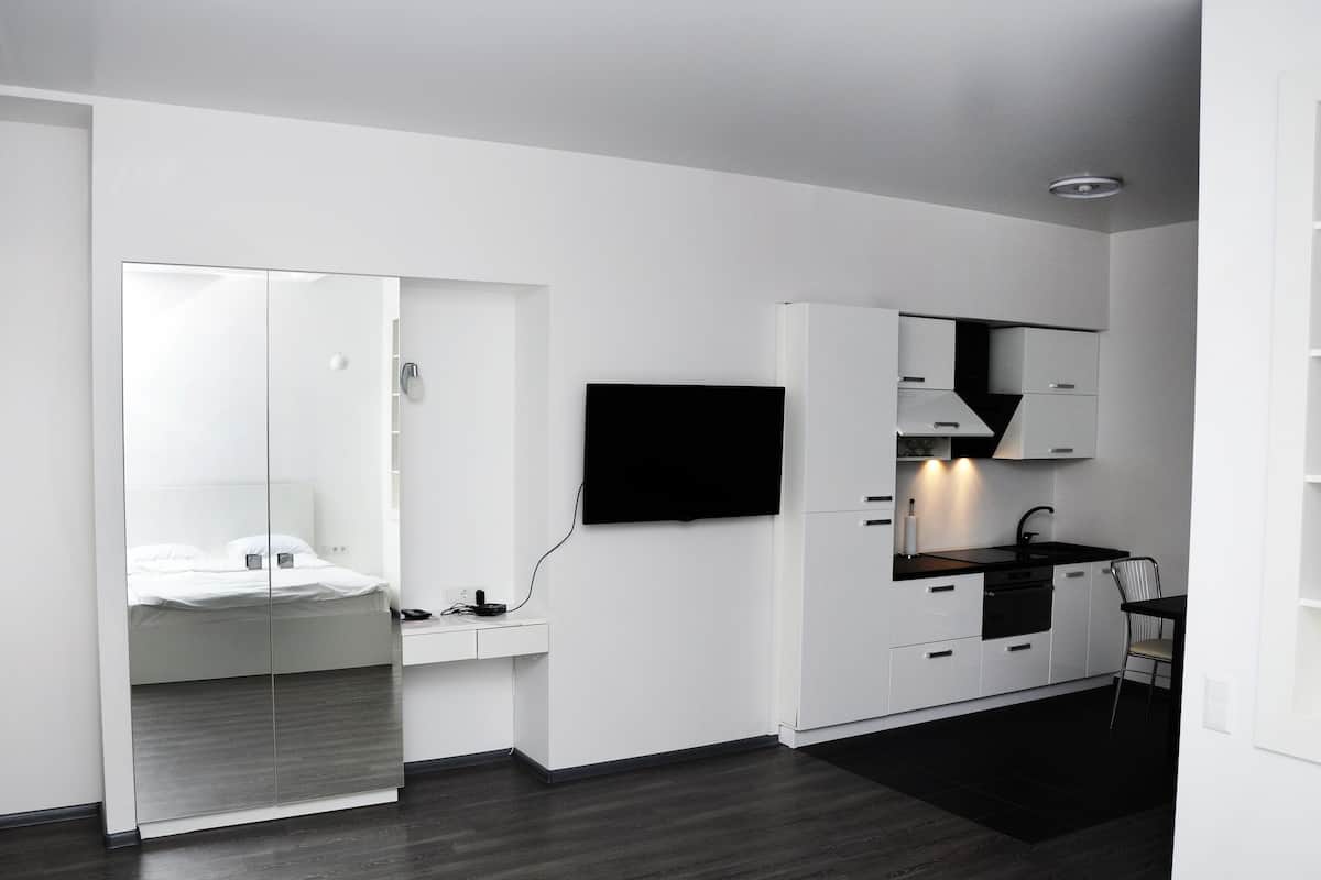 A modern, minimalistic room with a bed, a desk, a flat-screen TV, and a kitchenette.
