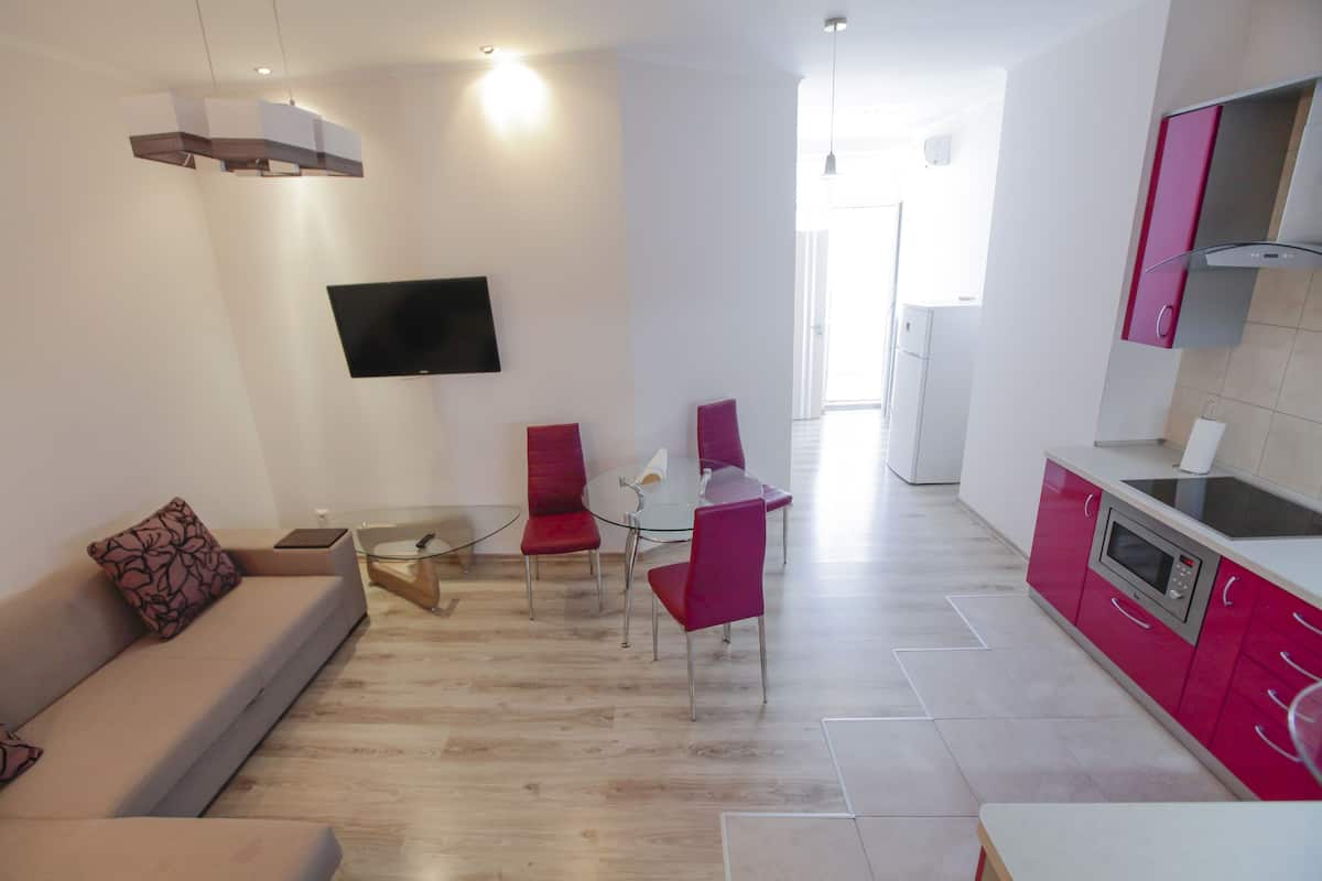 A modern living room with a sofa, a glass coffee table, and red chairs. There is a kitchen area with pink cabinets and a microwave.
