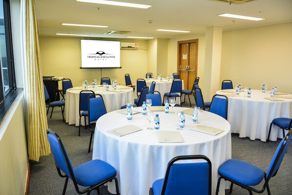 Un salón de conferencias con múltiples mesas redondas, cada una con sillas azules y manteles blancos. Hay botellas de agua y vasos sobre las mesas, y una pantalla de proyector que muestra el logotipo de Tropical Executive Hotel.