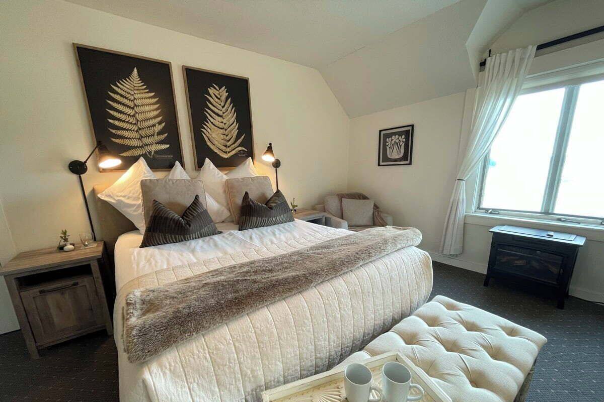 Un dormitorio ordenado con una cama grande, mesitas de noche, una lámpara de noche, una ventana con cortinas y una pequeña mesa con tazas.