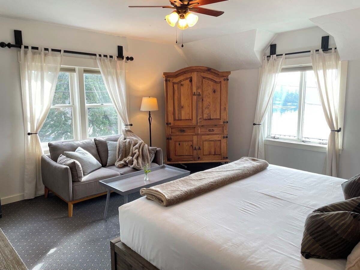 A bedroom with a bed, a sofa, a wooden wardrobe, a ceiling fan, and a window with curtains.