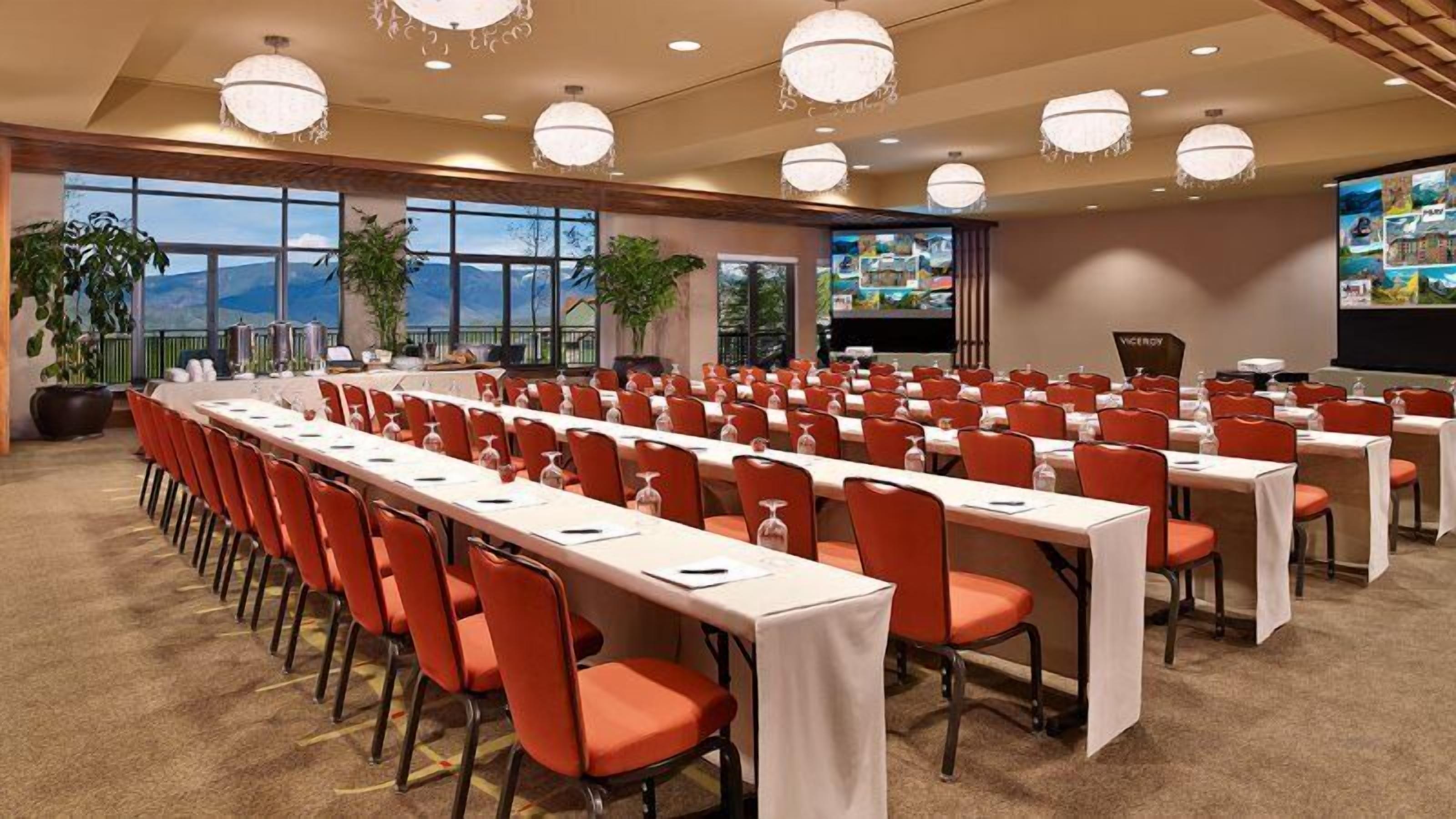 A conference room with rows of tables and chairs, large windows with a view, and a screen displaying images.