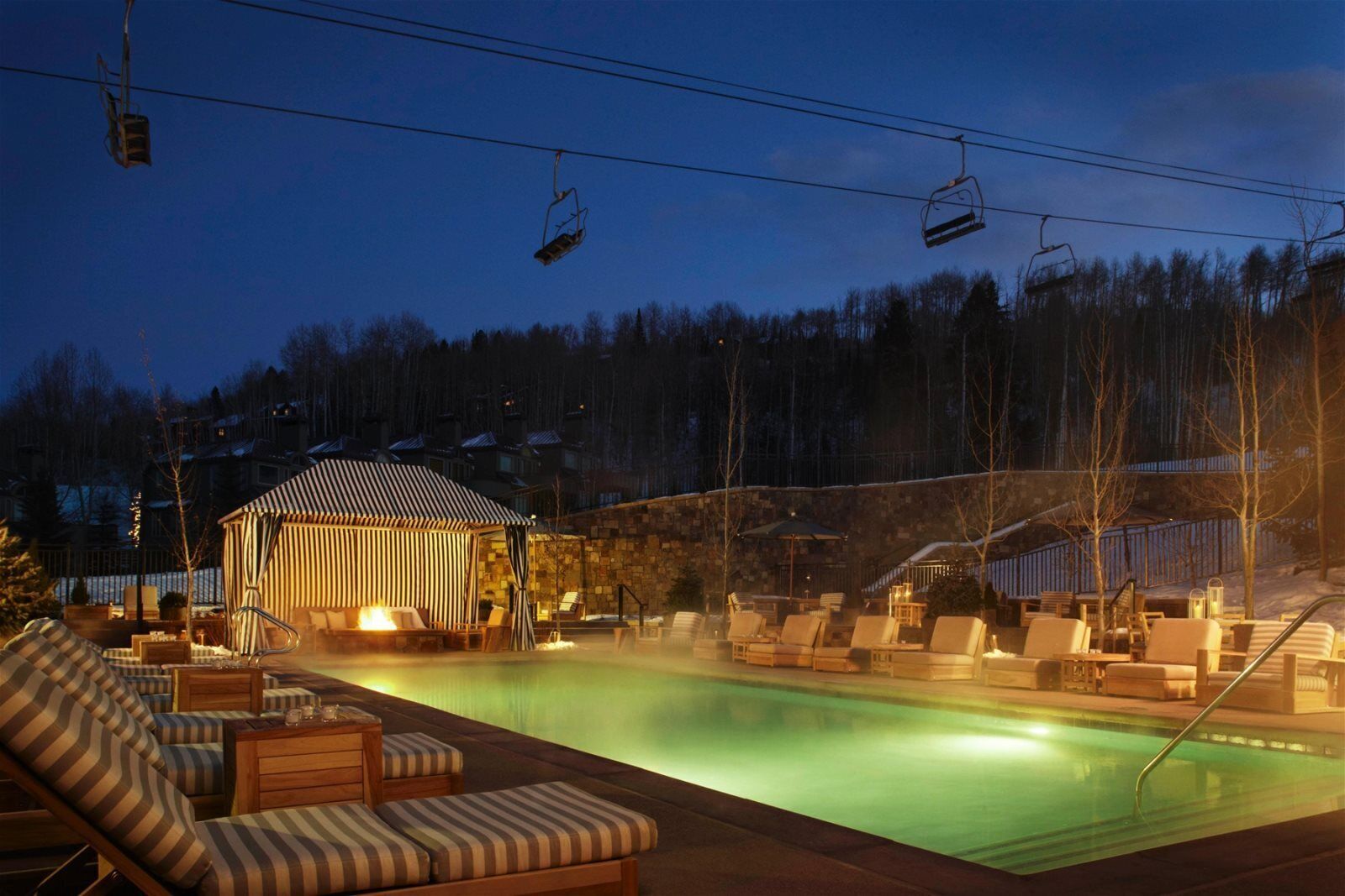 A heated outdoor pool area with lounge chairs and a covered seating area, surrounded by a stone wall and trees.