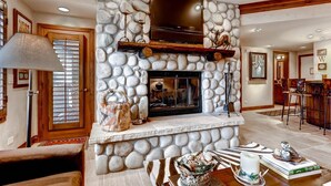 A stone fireplace with a wooden mantel, a television above it, and a dining area with a bar and chairs. - Beaver Creek Charter 2 Bed (Avon)