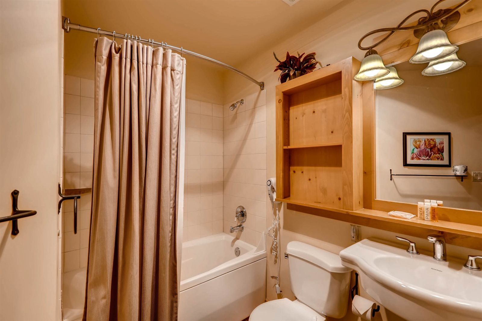 A bathroom with a shower curtain, a bathtub, a toilet, a sink, and a wooden shelf with toiletries.