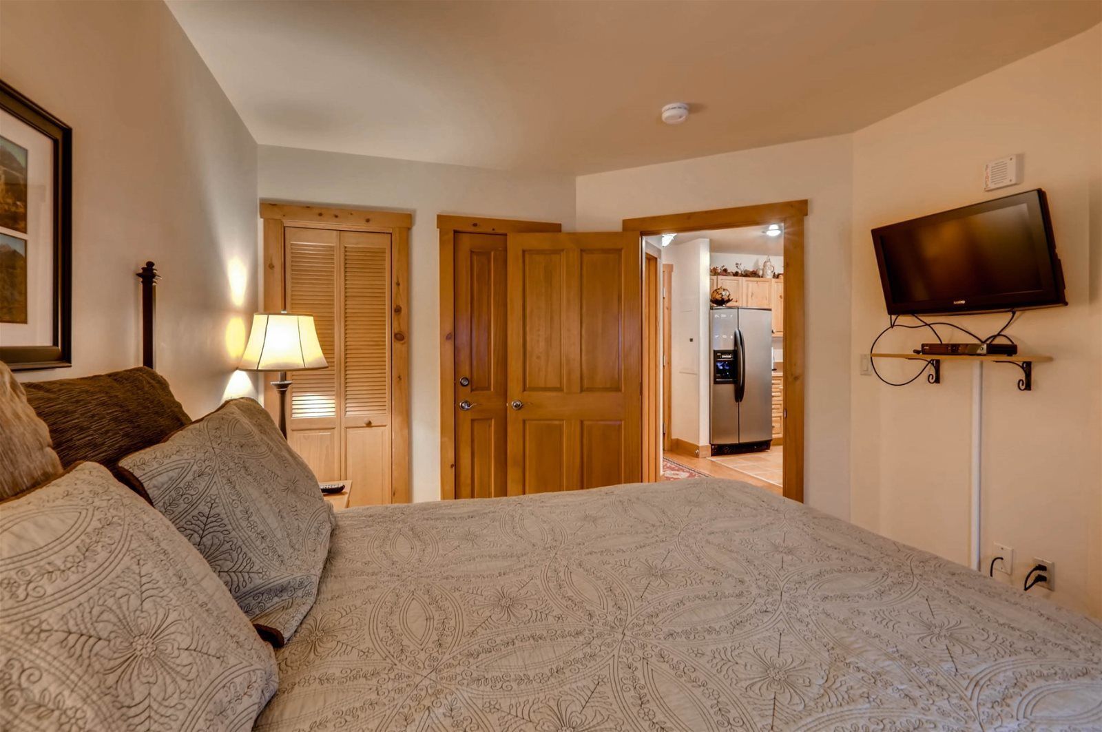 A bedroom with a large bed, wooden doors, a television mounted on the wall, and a kitchen visible through an open door.