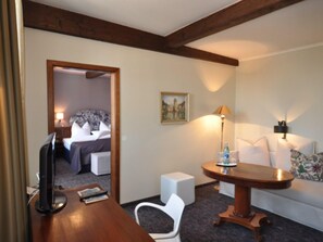 A hotel room with a wooden desk, a television, a small table with a lamp, a white chair, and a bed with a floral bedspread. - Hotel Schmuckkästchen (Miltenberg)