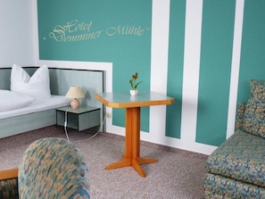 A hotel room with a bed, a nightstand, a small table with a plant, and a patterned armchair.