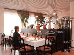 A dining area with a table set for two, a sign reading 'Hotel Demminer Mühle', and potted plants.