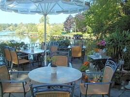 An outdoor patio with tables and chairs, surrounded by greenery and overlooking a body of water.
