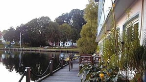 A wooden dock leading to a building with outdoor seating, overlooking a calm body of water and trees.