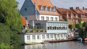 A riverside building with a prominent gabled roof, a balcony with seating, and a restaurant area with outdoor seating.