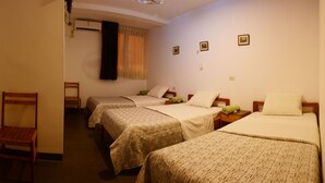 Quarto de hotel com três camas de solteiro, uma cadeira de madeira e uma pequena mesa com abajur.