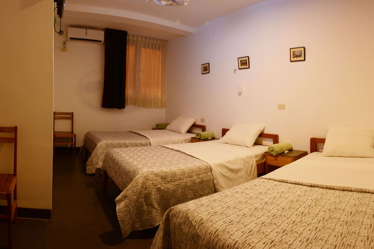 Ein Hotelzimmer mit drei Einzelbetten, einem Holzstuhl und einem kleinen Tisch mit einer Lampe.