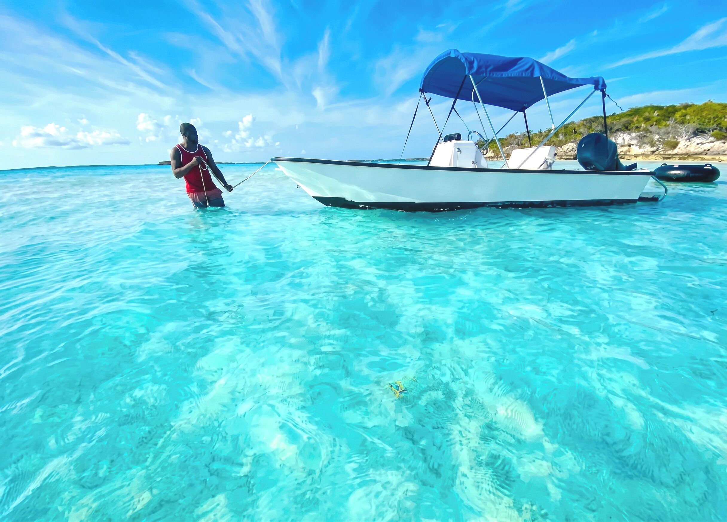 A boat anchored in clear turquoise water with a person standing on the shore.