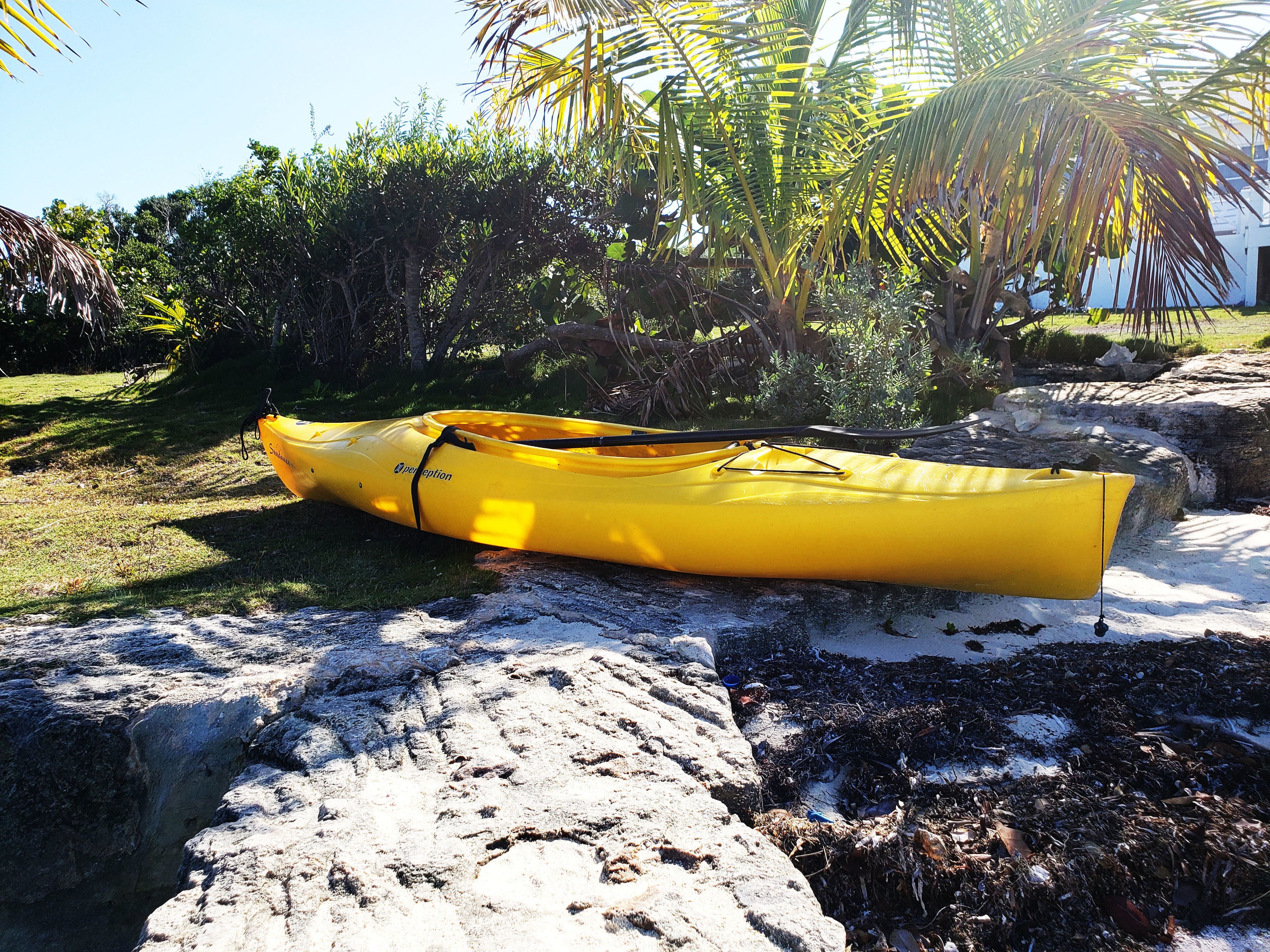 A yellow kayak resting on a rocky surface with greenery and a building in the background.