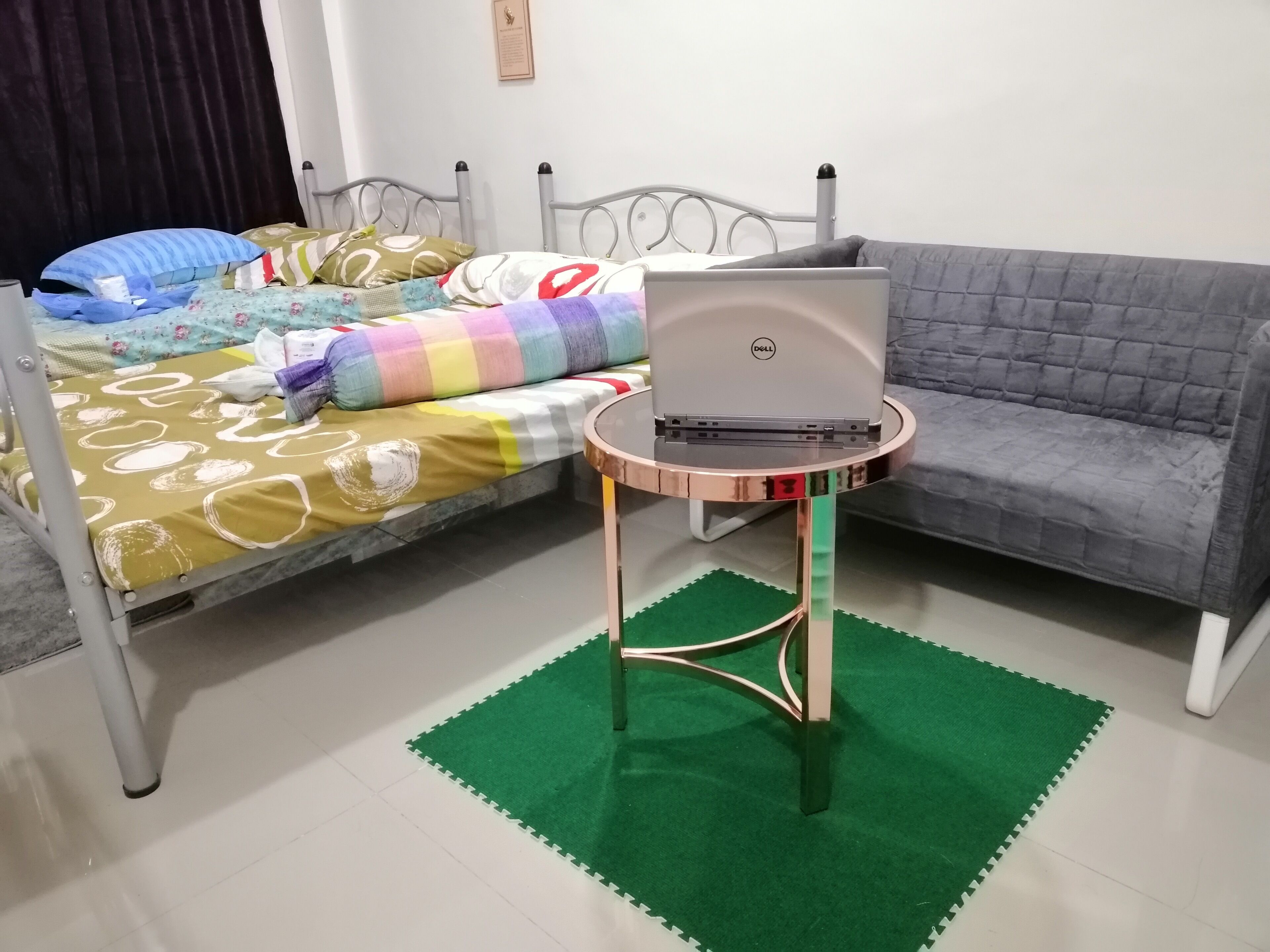 Une chambre à coucher avec un lit, un canapé gris, un ordinateur portable Dell posé sur une petite table et un tapis vert sur le sol.