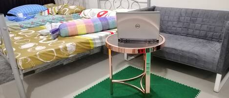 Une chambre à coucher avec un lit, un canapé gris, un ordinateur portable Dell posé sur une petite table et un tapis vert sur le sol.