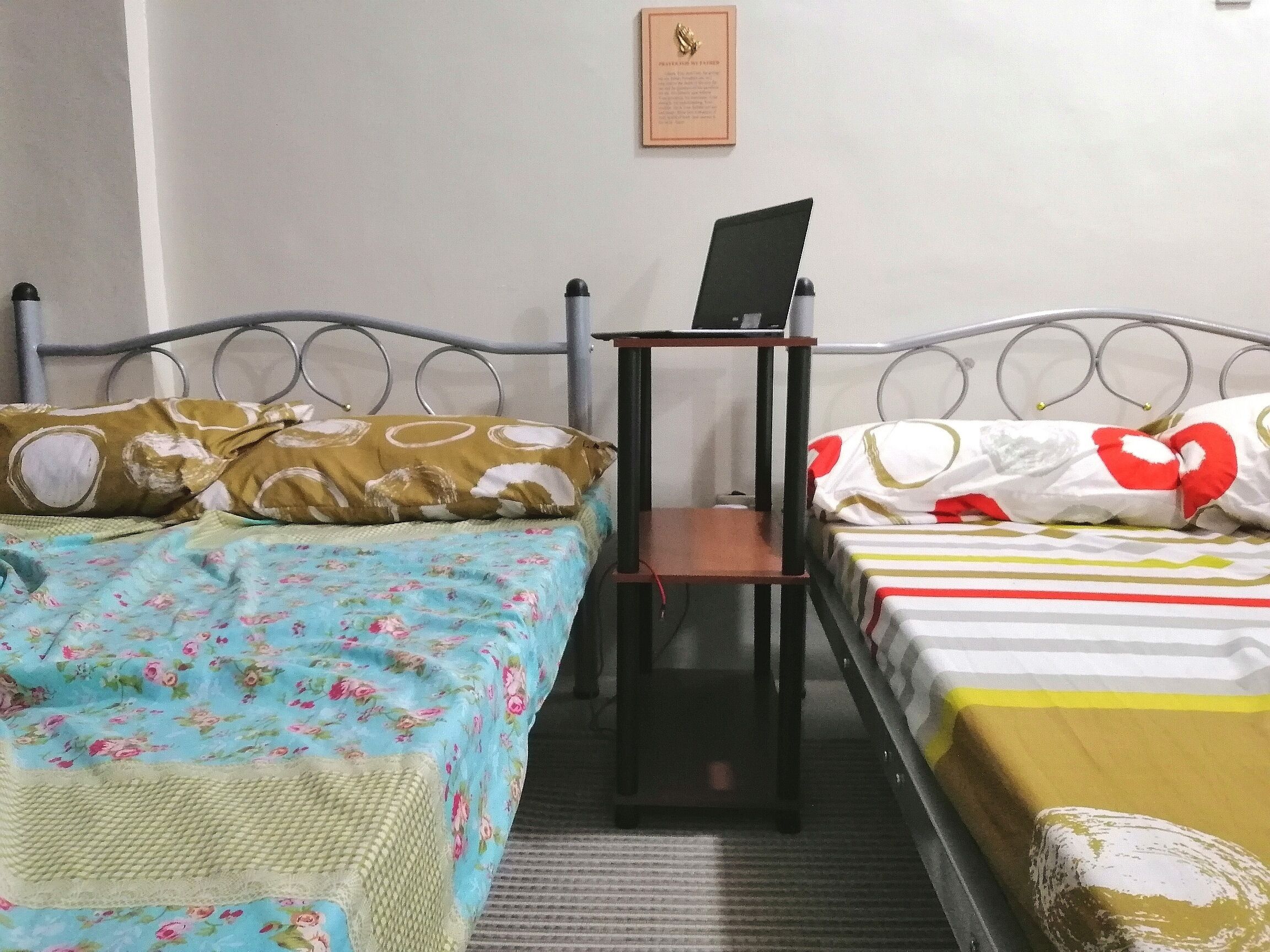 A room with two single beds, each with a distinct patterned bedspread. A small wooden table with a laptop is placed between the beds.
