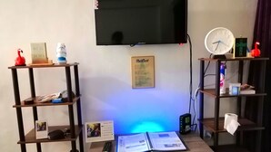 Una habitación con televisión de pantalla plana, un escritorio con un calendario, un control remoto y un libro. Estantes con diversos objetos a ambos lados.