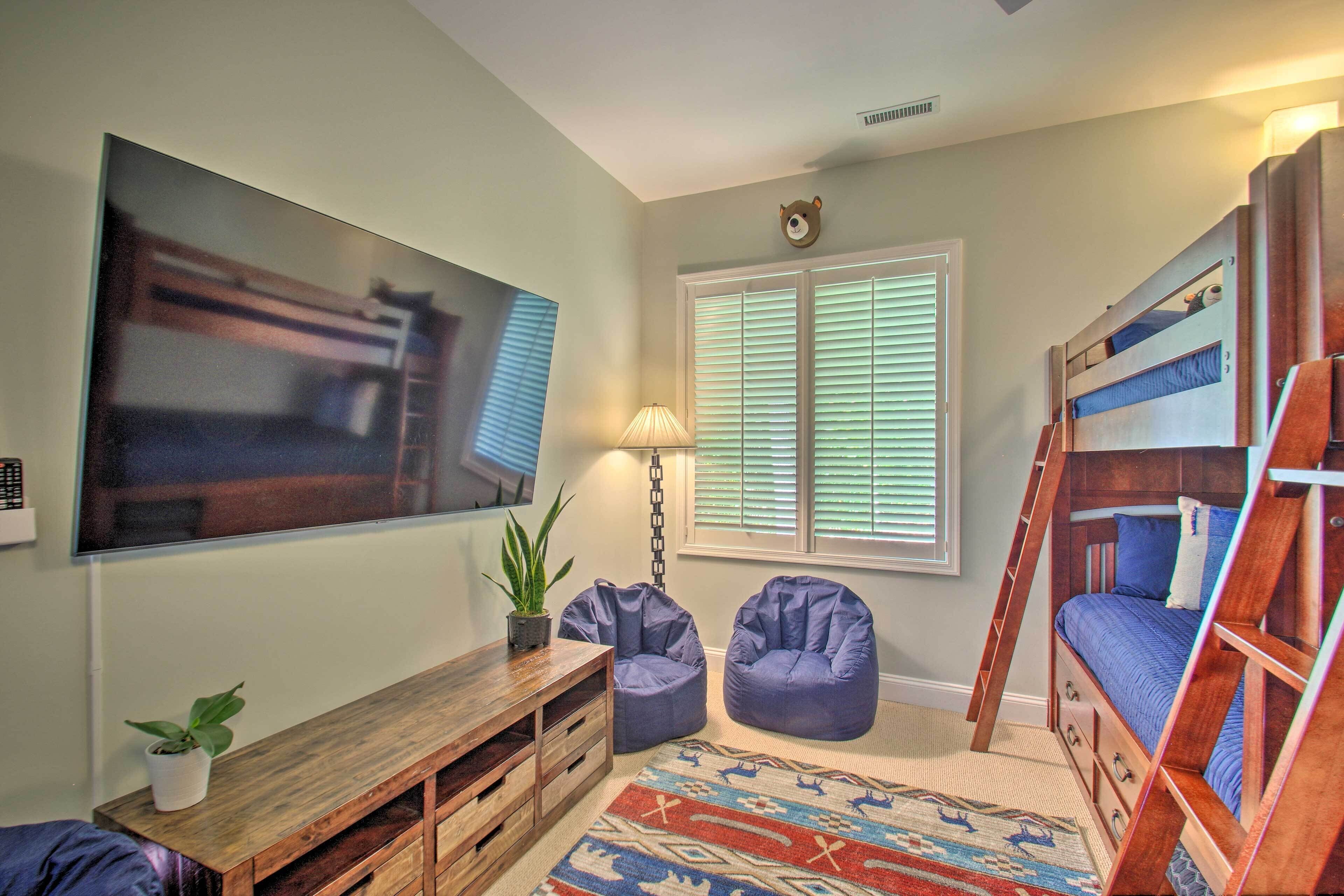 A room with bunk beds, a large TV, a wooden bench, two bean bags, a patterned rug, and a window with blinds.