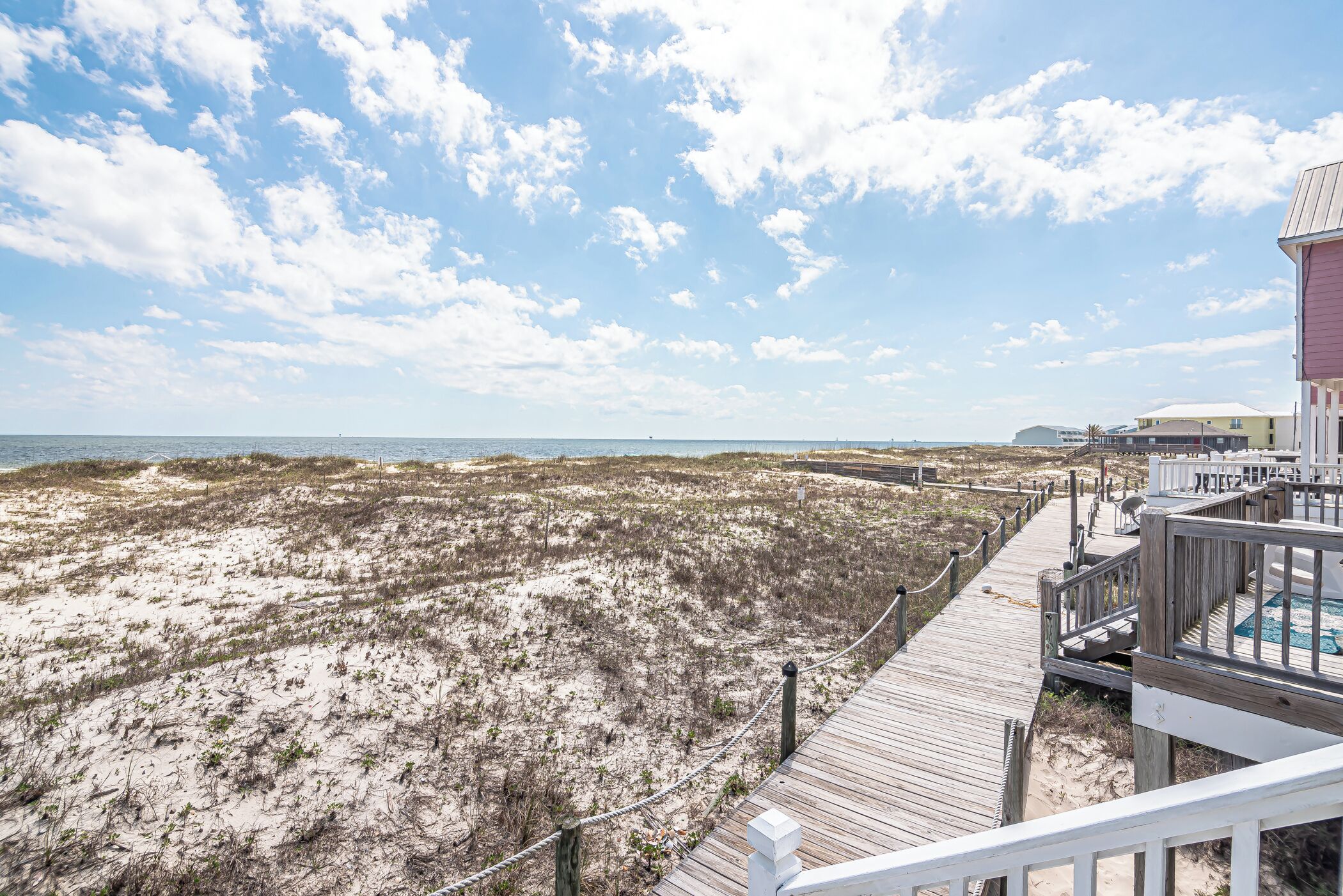 A beach view with a wooden boardwalk leading to a building with a pool.