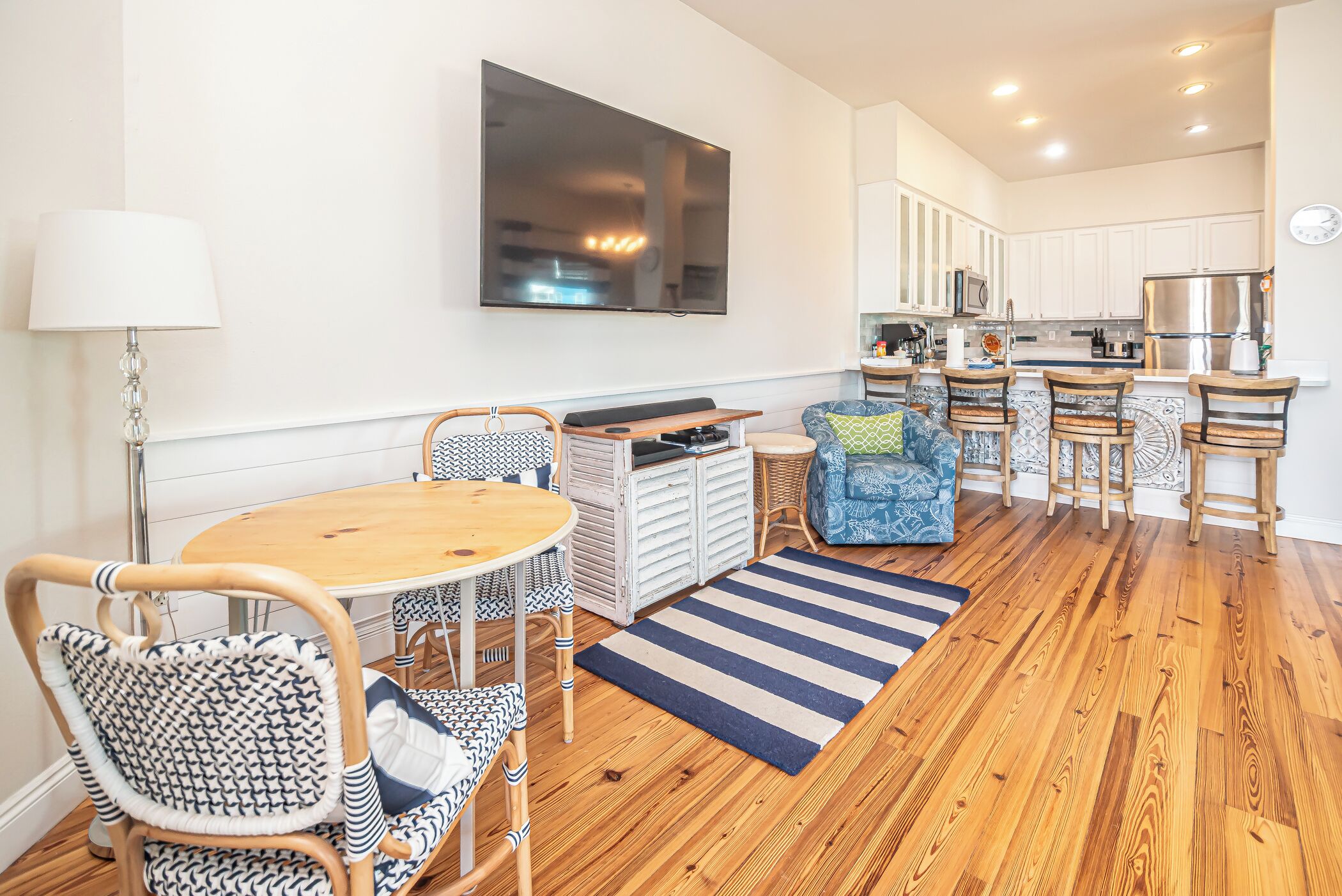 A modern kitchen with white cabinets, a large flat-screen TV, and a dining area with a wooden table and patterned chairs.