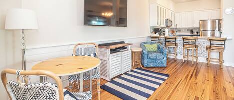 A modern kitchen with white cabinets, a large flat-screen TV, and a dining area with a wooden table and patterned chairs.