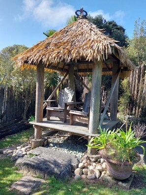 A wooden gazebo with a thatched roof, seating area, and a potted plant nearby. - Magical West Coast Retreat (Bethells Beach)