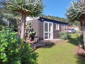A single-story house with a front porch, surrounded by a well-maintained lawn and various plants. - Magical West Coast Retreat (Bethells Beach)