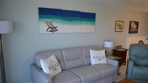 A living room with a grey sofa, two pillows with 'RELAX' text, a beach-themed painting, and a blue armchair. - Beachcomber unit 504 (Longboat Key)