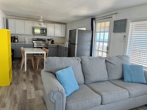A compact living space with a kitchenette, a gray sofa with blue cushions, and a view of the beach through the window. - Sun, surf , repeat ! On the beach ! Wake up to the waves!  (Surfside Beach)