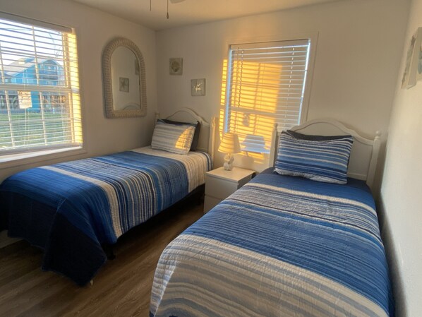 Une chambre avec deux lits simples, une table de chevet avec une lampe, un miroir et une fenêtre avec des stores.