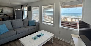 A living room with a gray sofa, a white coffee table, and a TV. There are three windows with blinds, one of which has a view of the ocean.