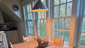 Un comedor con mesa de madera, sillas blancas, una lámpara colgante y vista a árboles a través de ventanas amplias.