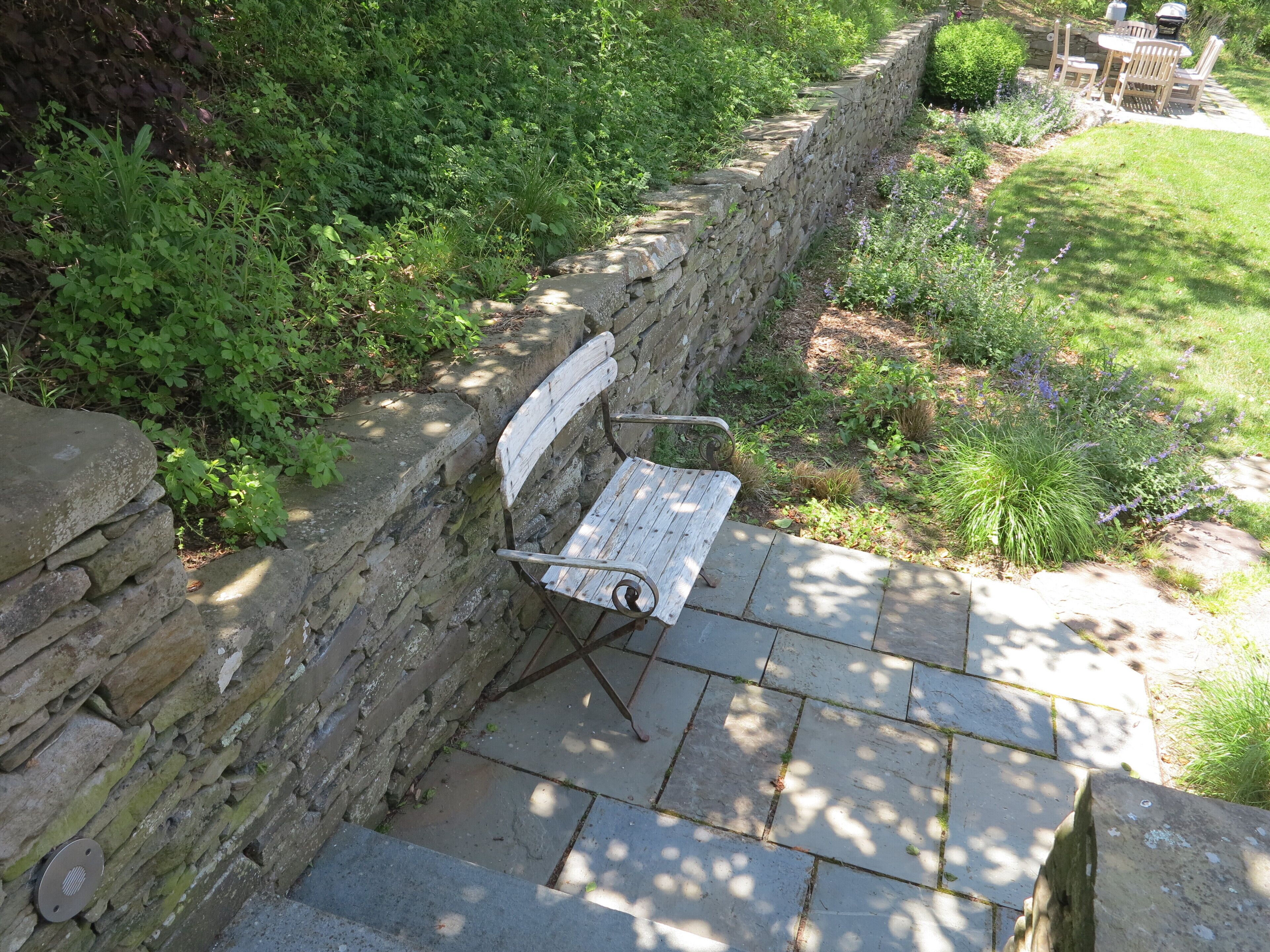 Eine Steinsitzbank mit Metallstützen, eine Steinmauer und eine gepflasterte Fläche mit Gras und Pflanzen.
